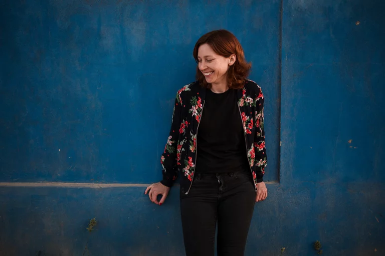 Eine Frau mit einer Blumenjacke steht mit dem Rücken an einer blauen Wand und blickt lachend von ihr aus gesehen nach rechts.
