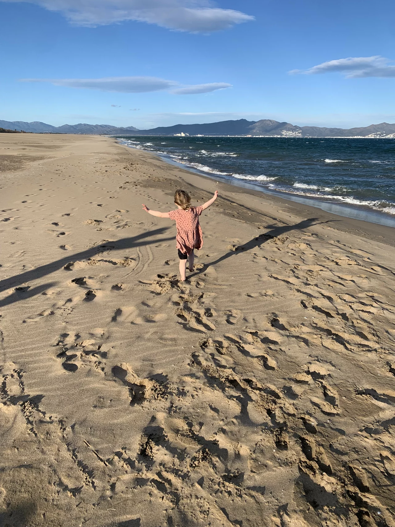 Ein Strand, auf dem ein kleines Kind in einem Kleid herumhüpft, man sieht es von hinten, wie es beide Arme in die Luft wirft und auf einem Bein hüpft.